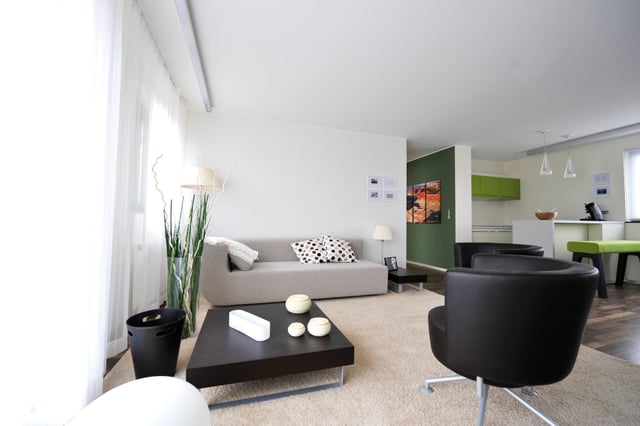 Wohnzimmer mit hellem Sofa, schwarzen Sesseln und dunklem Couchtisch, dahinter eine Küche