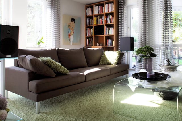 Braunes Sofa mit grünen Kissen vor einem Bücherregal und großen Fenstern