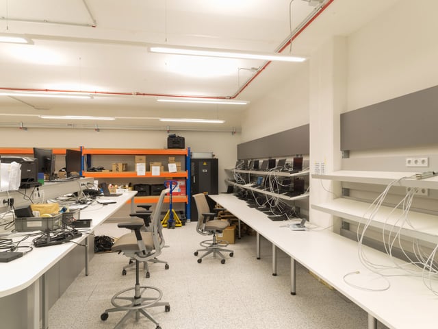 Raum mit langen weißen Arbeitstischen, Laptops auf Wandregalen und Drehstühlen