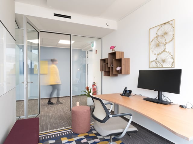 Einzelbüro mit geschwungenem Schreibtisch, Bildschirm und Wandregal, hinter der Glastür geht eine Person vorbei