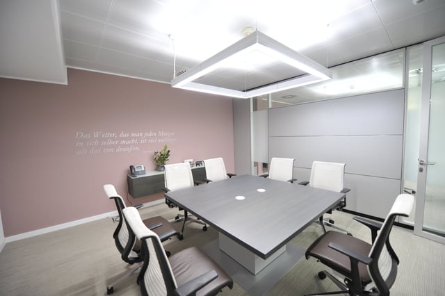 Besprechungsraum mit großem Tisch, weißen Drehstühlen und Wandspruch an rosa Wand
