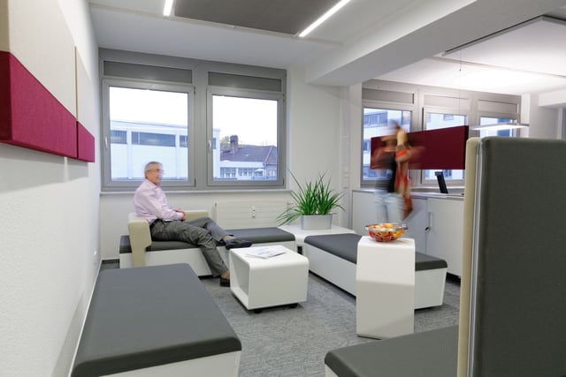 Lounge mit grauen Sitzbänken, Sitzwürfeln und roten Wandpaneelen, ein Mann sitzt auf einer Bank