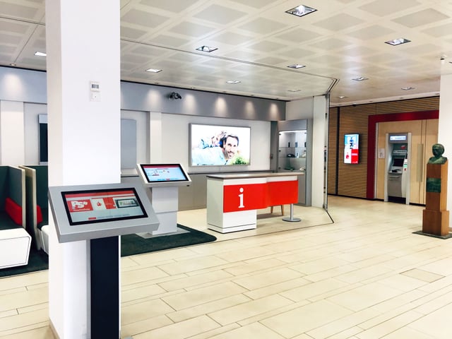 Kundenhalle mit Terminals, roter Infotheke und Geldautomat im Hintergrund