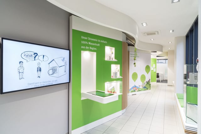 Kundencenter mit Bildschirm und grüner Wand mit dem Text „Unser Strommix ist einfach: 100% Wasserkraft aus der Region.“