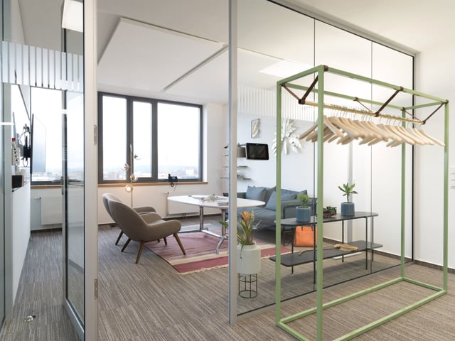 Lounge hinter Glaswänden mit Sesseln, Tisch und einer grünen Garderobe