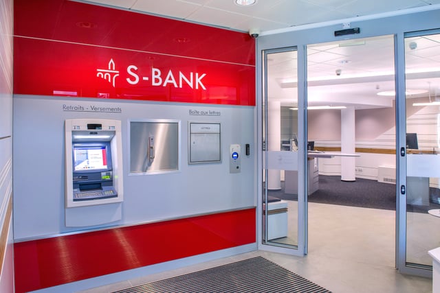 Selbstbedienungsbereich mit Geldautomat und dem Schriftzug „S-Bank“ auf roter Wand neben einer Glastür