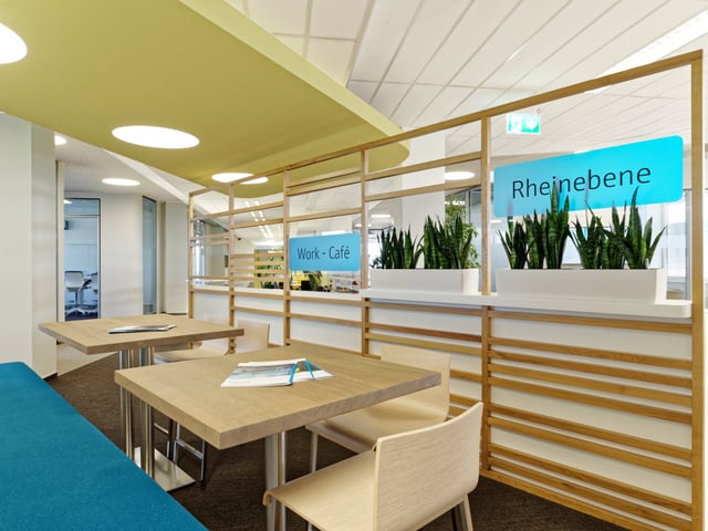 Sitzplatz mit Holztischen und blauer Bank hinter einer Holzlattenwand mit Pflanzen und den Schildern „Work - Café“ und „Rheinebene“