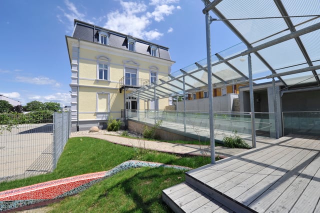 Gelbe Villa mit Rampe unter einem Glasdach, Holzterrasse und Rasen