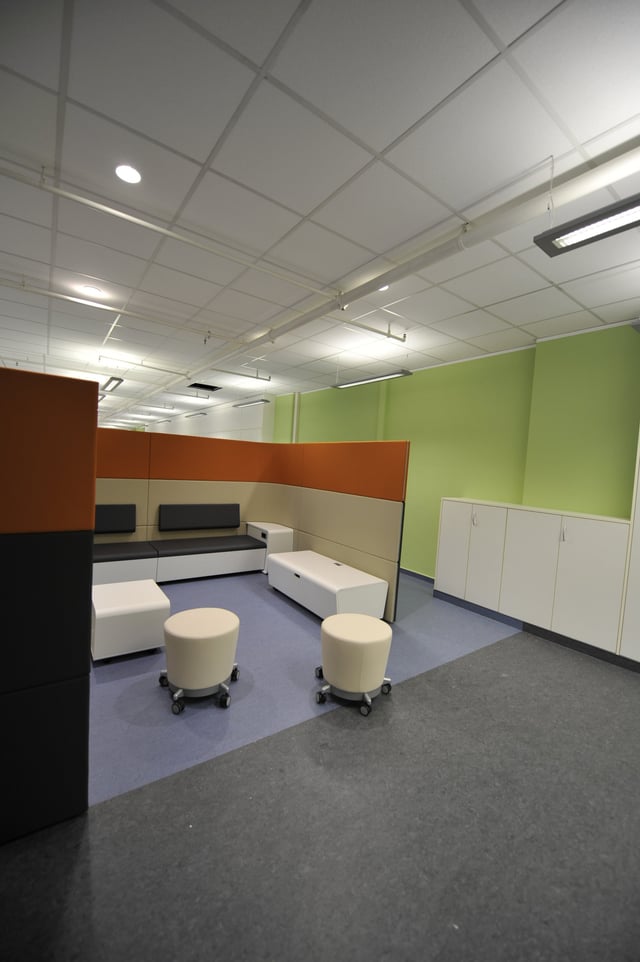 Sitzecke mit Bänken, orangen Rückwänden, Hockern und weißen Sitzwürfeln vor einer grünen Wand