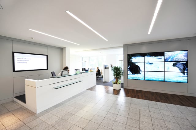 Empfangshalle mit weißer Theke, Bildschirm und Videowand, an der Theke sitzt eine Person