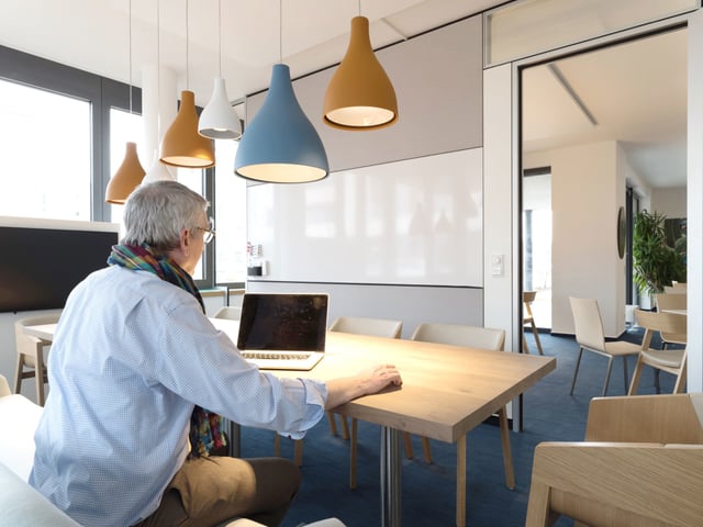 Mann mit Laptop an einem langen Holztisch unter farbigen Pendelleuchten