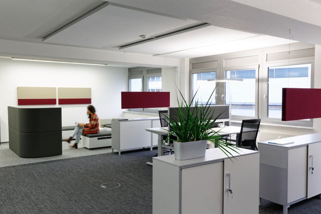 Bürofläche mit Schreibtischen und roten Tischtrennwänden, hinten eine Frau in einer Sitznische