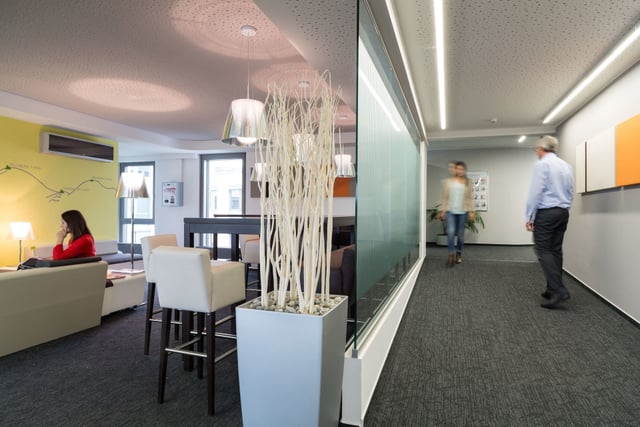 Lounge mit Sofas und Barhockern neben einem Flur mit Glaswand, eine Frau telefoniert