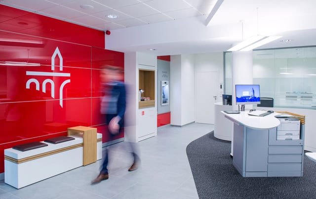 Kundenhalle mit roter Wand mit Logo, Sitzbank und Empfangstheke, ein Mann geht vorbei